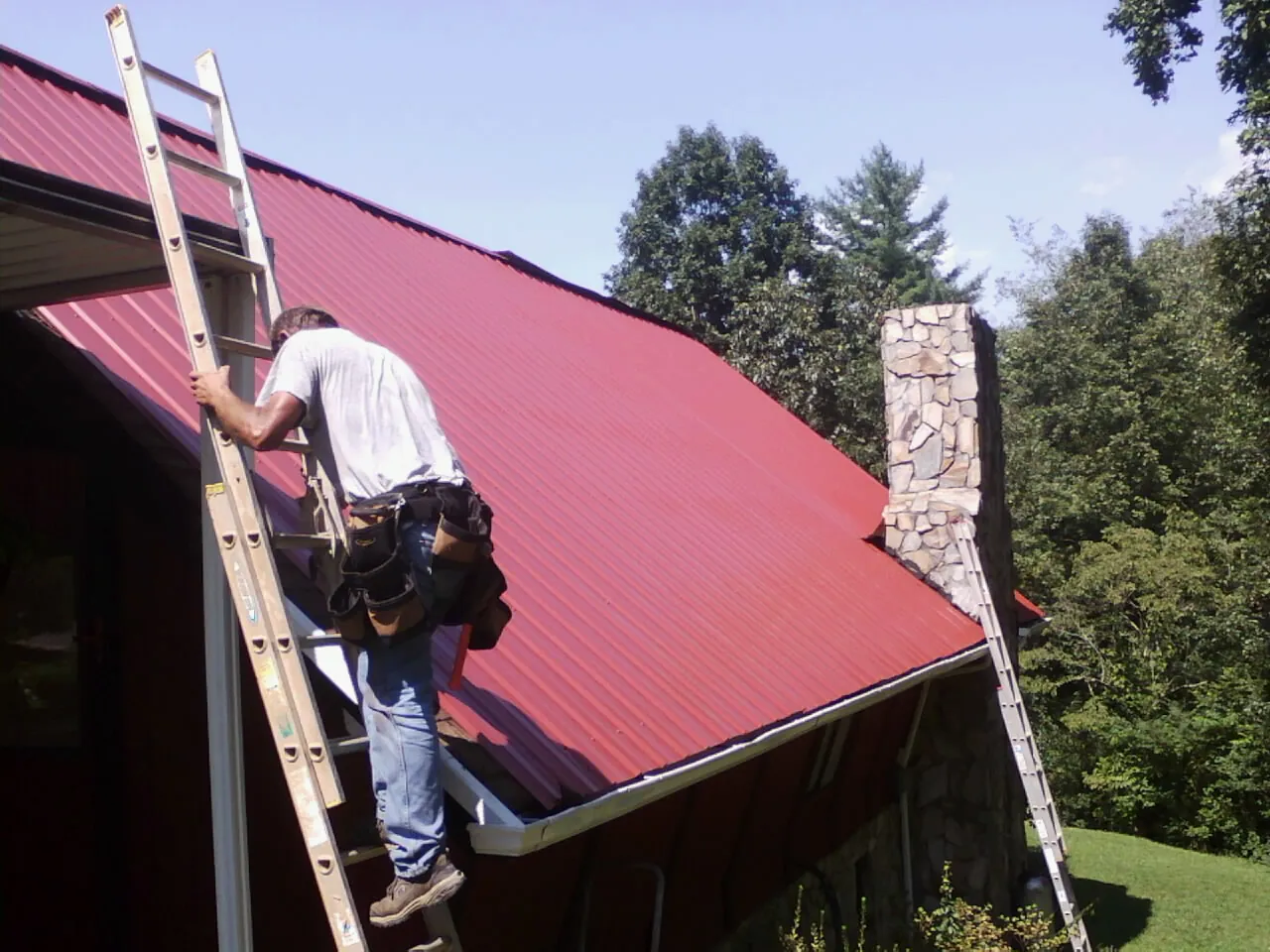Roofer on a ladder beside a red metal roof and stone chimney — Soffit & Fascia Repair by Roofing Brothers in Clarkdale